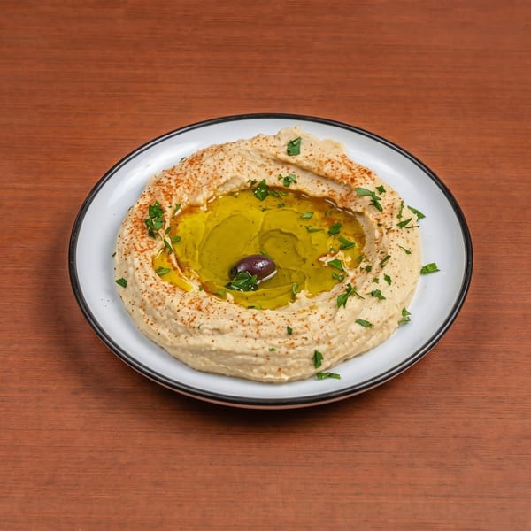 Creamy hummus with a pool of olive oil, paprika, parsley, and a single olive on a white plate.