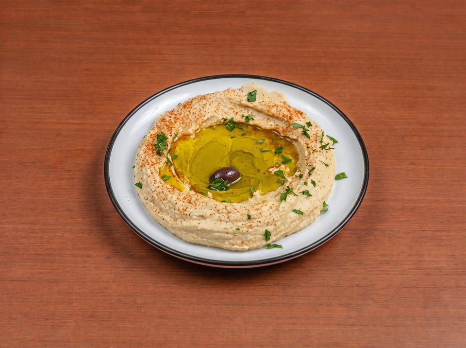 Creamy hummus with a pool of olive oil, paprika, parsley, and a single olive on a white plate.
