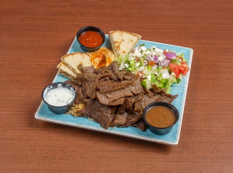 A gyro platter with sliced meat, rice, salad, pita bread, hummus, and sauces on a blue plate.