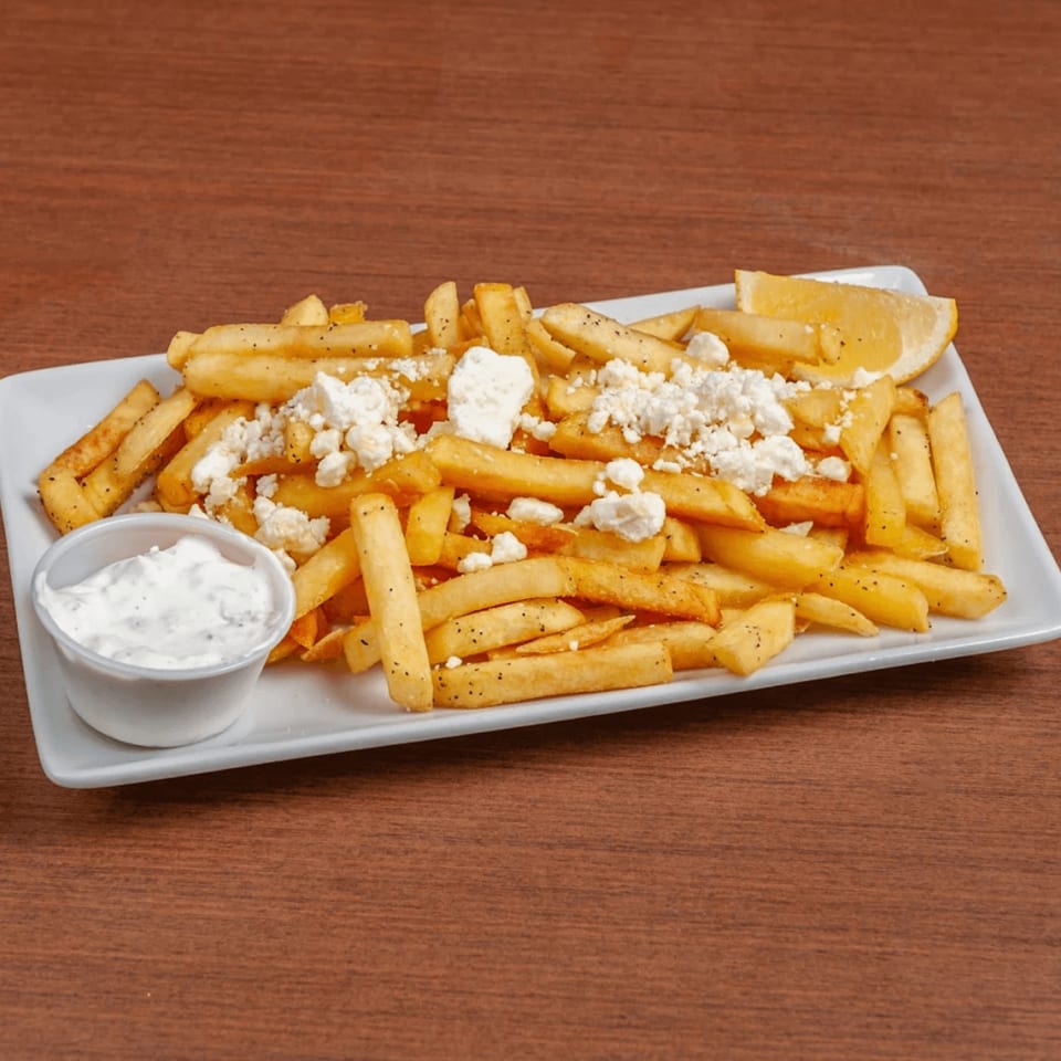 Golden french fries topped with crumbled feta cheese, black pepper, a lemon wedge, and a side of tzatziki sauce.