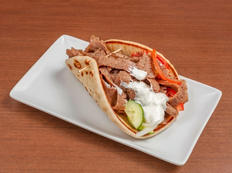 A gyro sandwich with sliced meat, vegetables, and white sauce on a white plate.