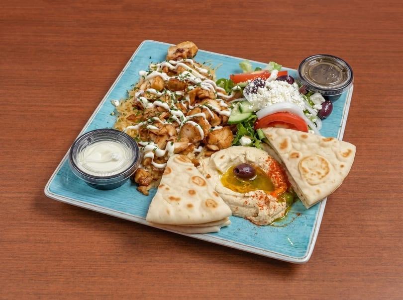 Chicken shawarma plate with rice, Greek salad, hummus, pita bread, and sauces on a blue plate.