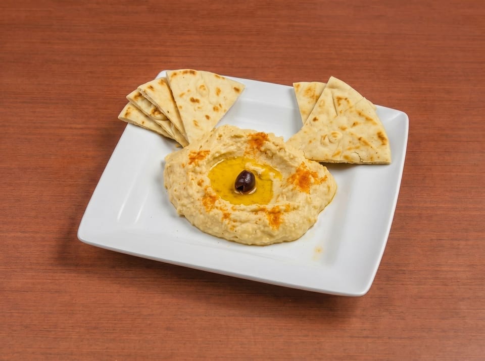 Creamy hummus with olive oil, a black olive, paprika, and pita bread on a white plate.